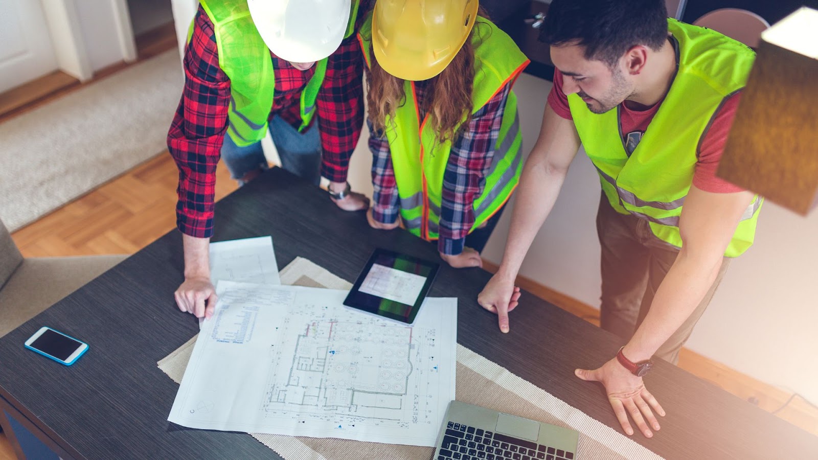 Three people in hard hats and safety vests looking at blueprints