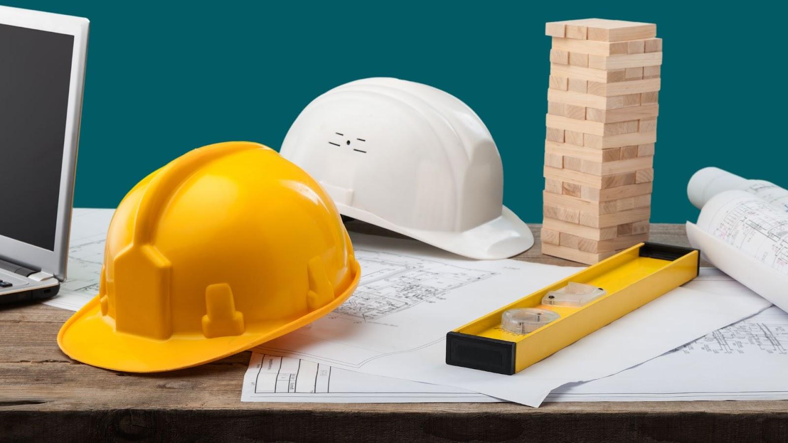 A laptop, construction tools and a hard hat on a table. 