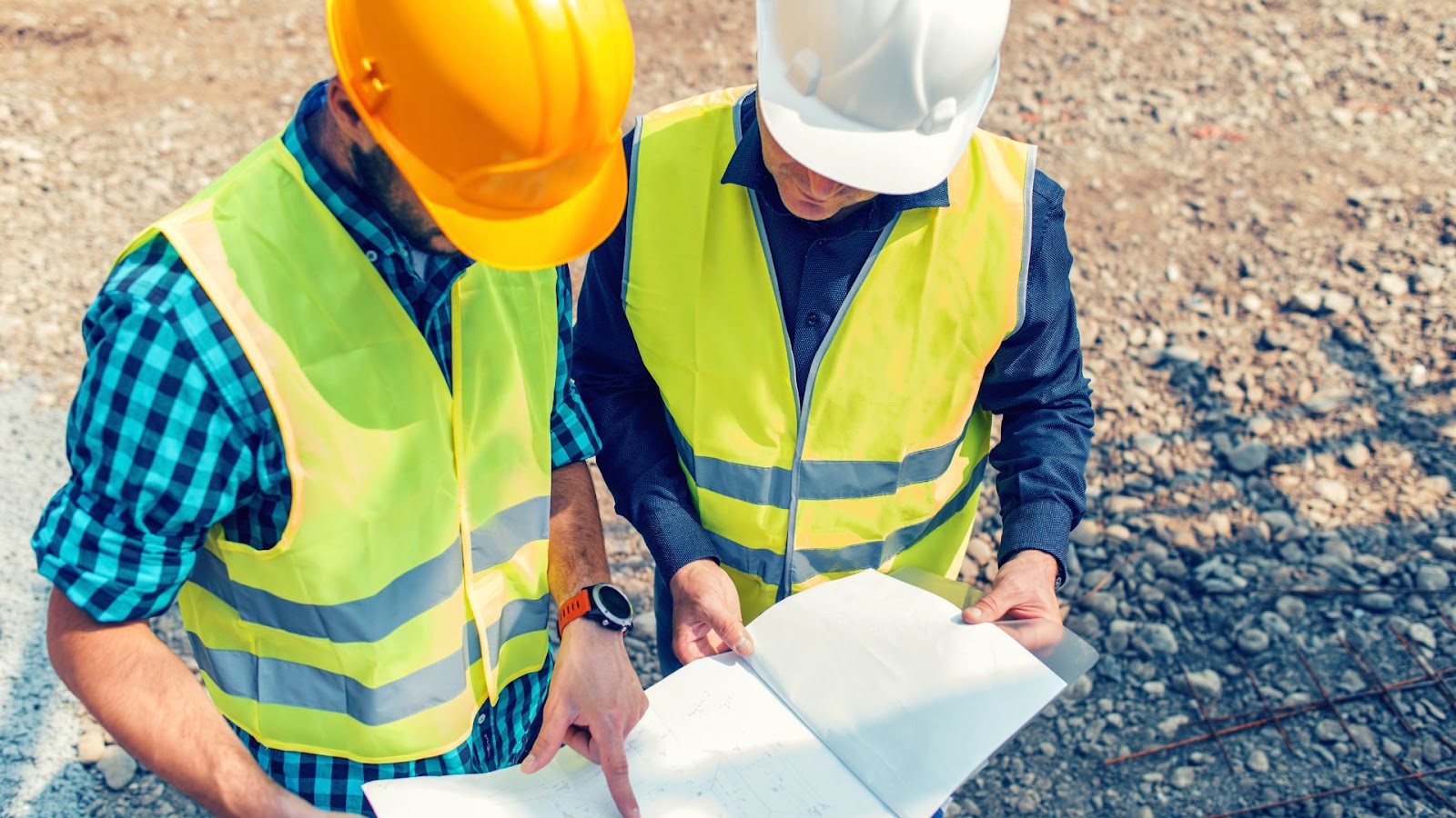 Two individuals in hard hats and safety vests looking at blueprints