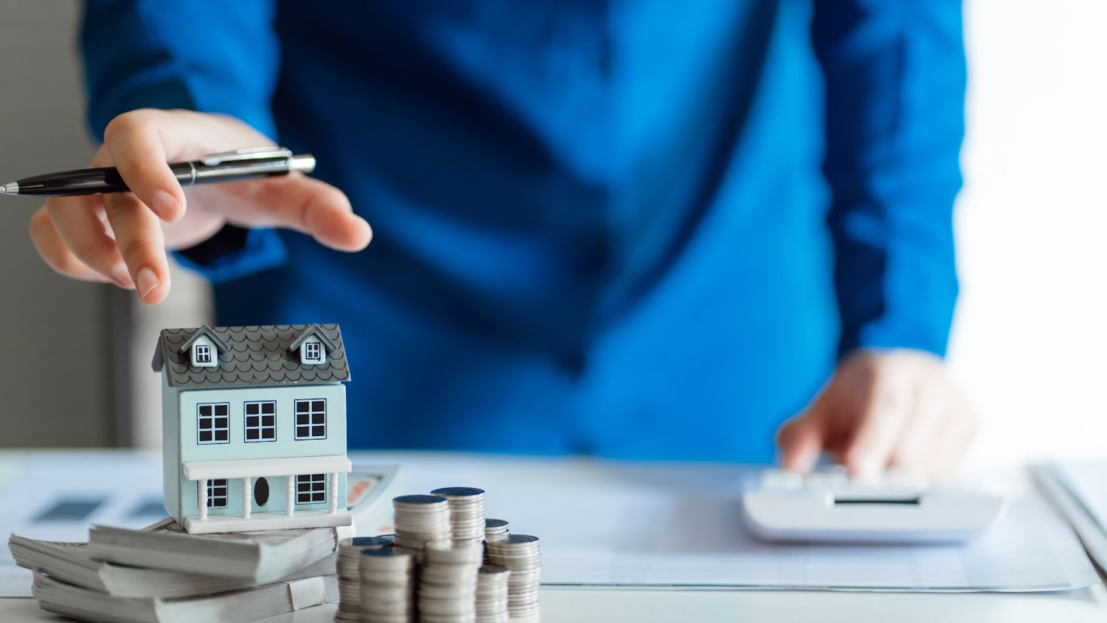 A person is holding a house model on top of a stack of coins