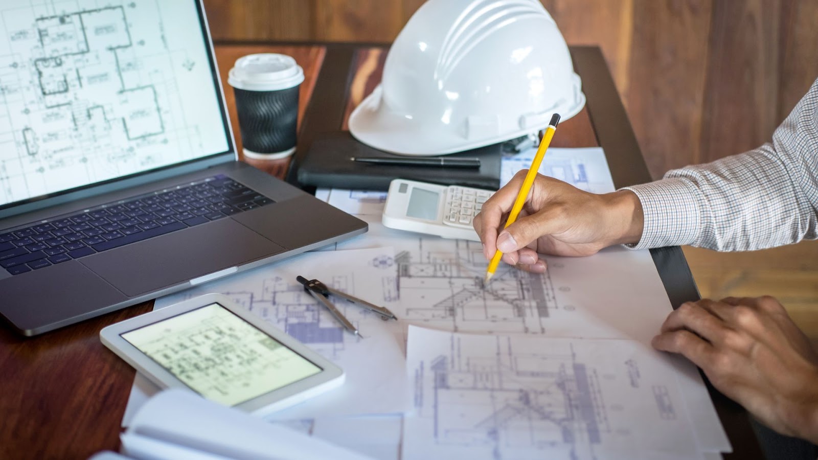 Person is working on a laptop with papers, a hard hat and coffee cup on table